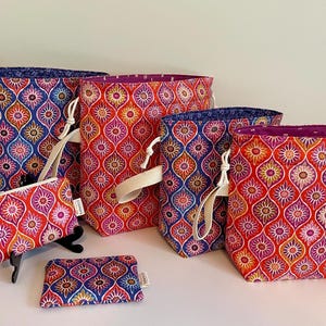 Puede incluir: Una colección de bolsas y estuches de tela con un vibrante estampado floral en rojo, azul y amarillo. El conjunto incluye bolsas con cordón, un bolso de hombro y pequeños estuches con cremallera. Las bolsas tienen cordones y correas blancos.
