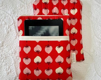Pode incluir: Duas capas de tecido vermelho com um padrão de morangos. Uma capa está aberta, segurando um e-reader preto. A outra capa está fechada com um zíper. O padrão de morangos apresenta morangos rosa e brancos em um fundo vermelho.
