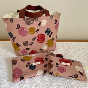 Może przedstawiać: Różowa torba i dwie pasujące saszetki z kolorowym nadrukiem jabłek. Torba ma brązowe uchwyty. Saszetki mają białą metkę z tekstem. Nadruk jabłek zawiera czerwone, żółte, brązowe i niebieskie jabłka.