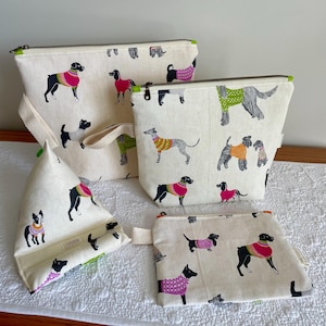 May include: A collection of cream-colored fabric bags and accessories, each adorned with a repeating pattern of dogs wearing colorful sweaters. The bags include zippered pouches, a tote bag, and a book stand, all with green zipper pulls.