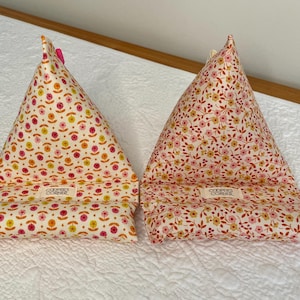 May include: Two triangular fabric phone stands. One is cream-colored with a floral pattern in orange, yellow, and pink. The other is pink with a similar floral design. Each has a label that reads "COOPER'S CORNER".