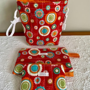 May include: A set of quilted fabric bags in a vibrant floral pattern. The set includes a bucket-style bag with white rope handles, and two zippered pouches. The bags feature a red background with colorful flower designs and the text "COOPER'S CORNER".
