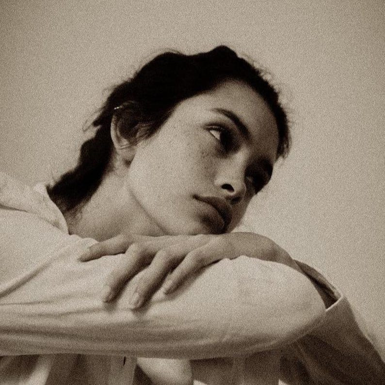 May include: Sepia-toned portrait of a person with dark hair and fair skin, resting their head on their arm. The person is looking off to the side, with a thoughtful expression. The image is softly lit, creating a vintage aesthetic.