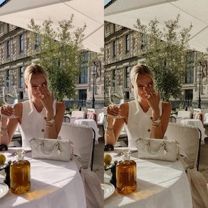 May include: A woman wearing a white sleeveless dress sits at a table outside a cafe. She is holding a glass of white wine and smiling. There is a white handbag on the table next to a bottle of olive oil.