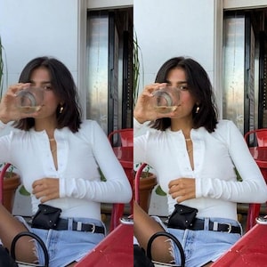 May include: A woman wearing a white long-sleeved shirt with a button-down front, denim shorts, and a black belt with a small black bag attached. She is holding a glass of white wine and has a gold chain necklace on.
