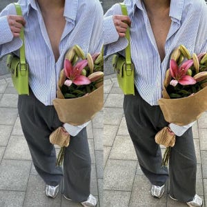 May include: A person wearing a light blue striped shirt with a green crossbody bag and gray pants. They are holding a bouquet of pink lilies wrapped in brown paper.