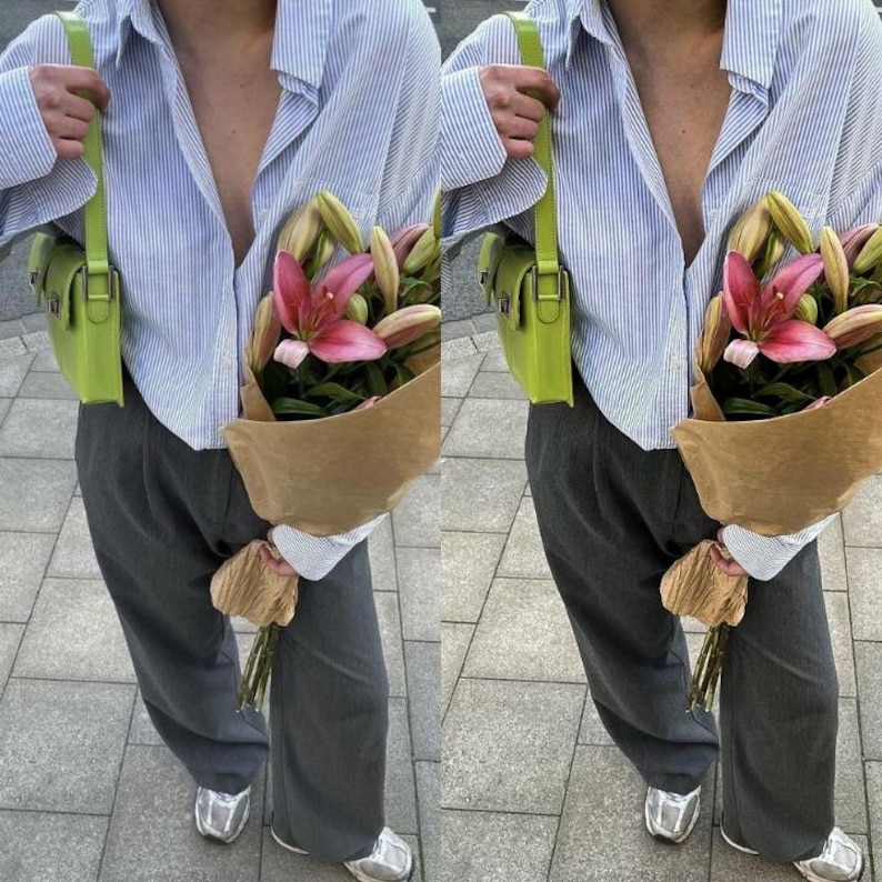 May include: A person wearing a light blue striped shirt with a green crossbody bag and gray pants. They are holding a bouquet of pink lilies wrapped in brown paper.