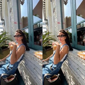 May include: A woman wearing a white tank top and blue denim overalls is sitting outside a cafe. She is holding a cup of iced coffee and wearing sunglasses.