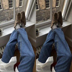 Puede incluir: Un par de zuecos de ante color topo con correa de hebilla lateral, usados con calcetines de canal&eacute; gris claro y pantalones vaqueros azules. El calzado presenta una puntera redondeada y una suela plana, ofreciendo un estilo informal.