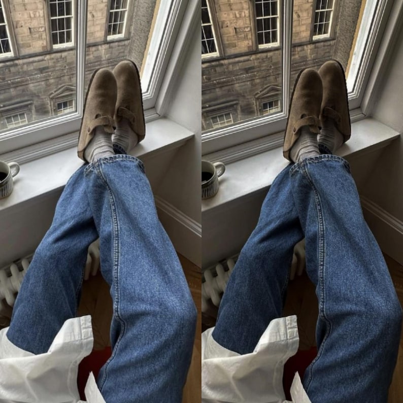 Puede incluir: Un par de zuecos de ante color topo con correa de hebilla lateral, usados con calcetines de canal&eacute; gris claro y pantalones vaqueros azules. El calzado presenta una puntera redondeada y una suela plana, ofreciendo un estilo informal.