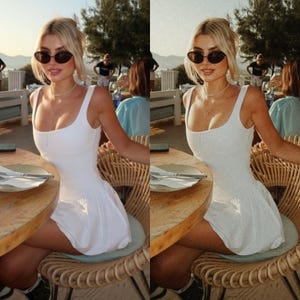 May include: A woman wearing a white sleeveless dress with a scoop neck and a fitted bodice. She is sitting on a wicker chair at a table with a white plate and silverware. She is wearing sunglasses and a gold necklace.