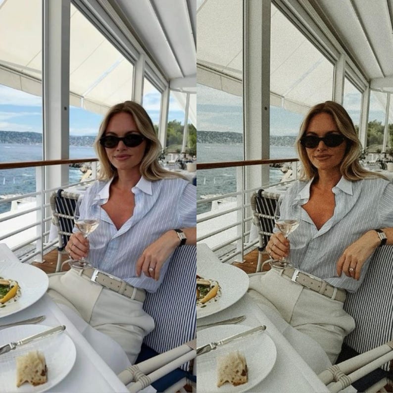 May include: A woman wearing a light blue striped button-down shirt with a white collar, white pants, and black sunglasses sits at a table with a glass of white wine. The table is set with a white tablecloth and a plate of food. The woman is sitting in a restaurant with a view of the water.