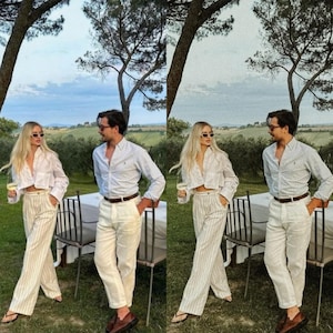May include: A couple wearing white shirts and striped pants stand in a garden setting. The man is wearing a brown belt and the woman is wearing sunglasses. They are both holding glasses of wine.