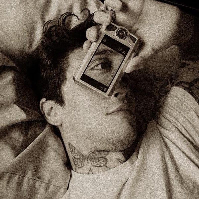 May include: A sepia-toned image of a person holding a digital camera over their eye. The camera's screen reflects the person's eye. A butterfly tattoo is visible on the person's neck.