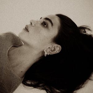 May include: A sepia-toned image of a person looking upwards. The person is wearing a light-colored, open-knit sweater and small hoop earrings. Dark hair is styled away from the face. The image is a close-up, focusing on the face and upper body.