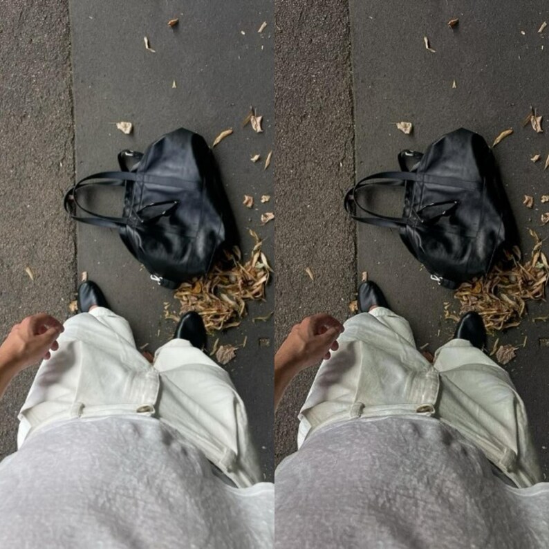 May include: A person wearing white pants and a grey shirt is standing on a sidewalk with a black leather tote bag and fallen leaves.