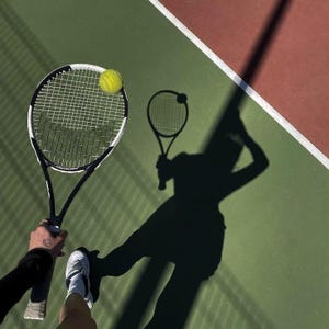 Puede incluir: Una raqueta de tenis negra y blanca con una pelota de tenis amarilla brillante sobre las cuerdas. La raqueta se sostiene sobre una superficie de pista de tenis verde y roja, proyectando una larga sombra oscura que muestra la silueta.