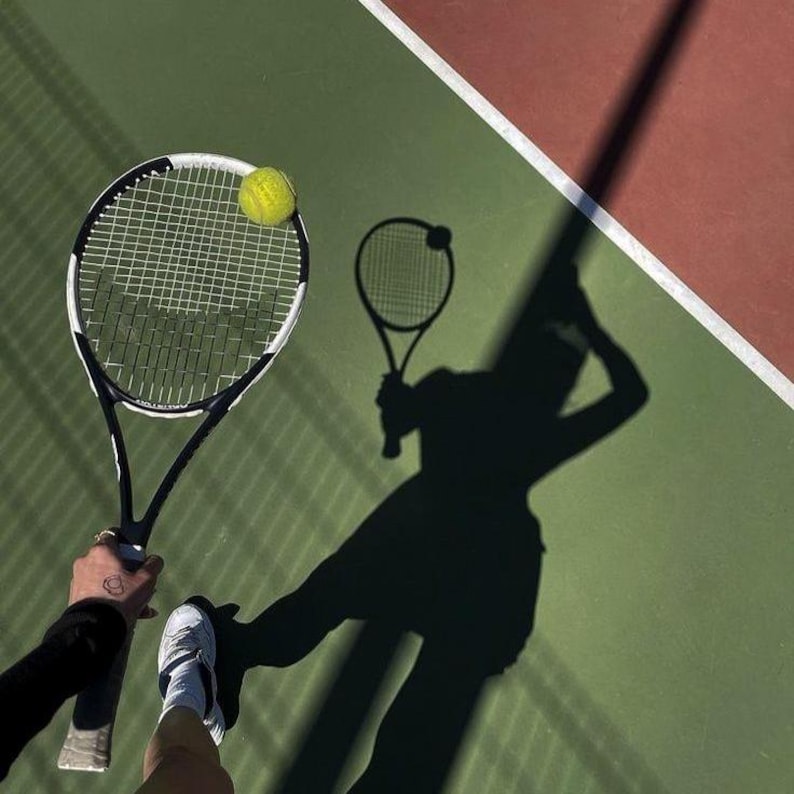 Puede incluir: Una raqueta de tenis negra y blanca con una pelota de tenis amarilla brillante sobre las cuerdas. La raqueta se sostiene sobre una superficie de pista de tenis verde y roja, proyectando una larga sombra oscura que muestra la silueta.