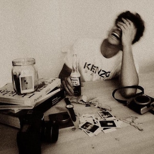 May include: A sepia-toned image featuring a table with a bottle of Corona beer, a jar, books, a watch, and a camera. A person wearing a white t-shirt with the word "KENZO" is laughing. Scattered photos and other items are on the table.