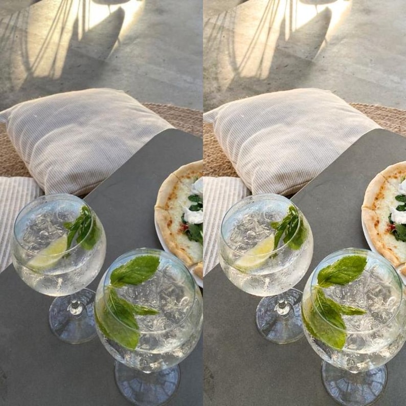May include: Two clear glass goblets filled with a refreshing drink, garnished with fresh mint and a slice of lime. The goblets are on a gray table next to a plate of pizza with a white crust and green toppings.