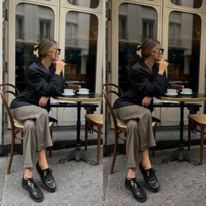 Puede incluir: Un blazer negro, pantalones de color marr&oacute;n claro y zapatos de cuero negro. El conjunto incluye una pinza para el pelo blanca y gafas de sol oscuras. La vestimenta se lleva puesta mientras se est&aacute; sentado en una mesa redonda peque&ntilde;a.