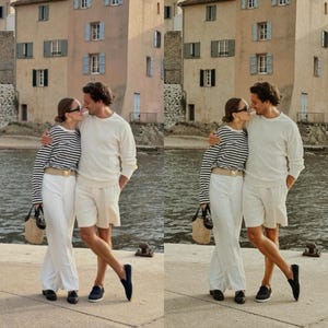 May include: A couple wearing white clothing and striped shirts are standing on a dock by the water. The man is wearing white shorts and a white sweater. The woman is wearing white pants and a black and white striped shirt. They are both wearing sunglasses.
