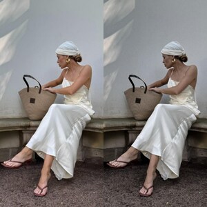 May include: A woman wearing a white turban and a white slip dress sits on a stone bench holding a brown woven tote bag. She is wearing brown heeled sandals.