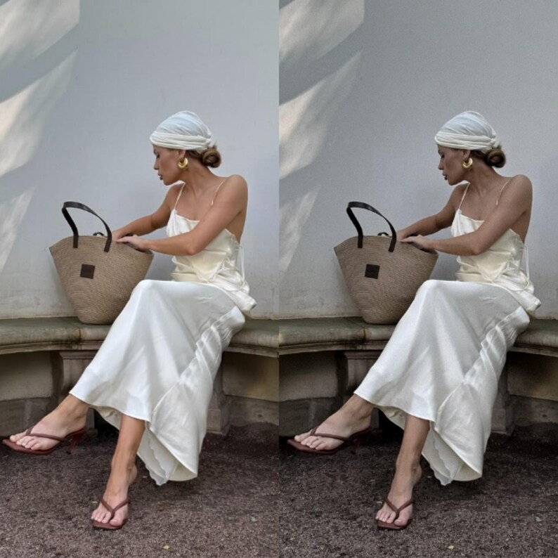 May include: A woman wearing a white turban and a white slip dress sits on a stone bench holding a brown woven tote bag. She is wearing brown heeled sandals.