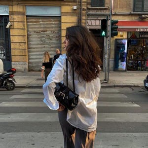 Puede incluir: Una camisa blanca de manga larga con botones, combinada con pantalones oscuros y un bolso de hombro de charol negro con una hebilla plateada prominente. La camisa tiene un corte relajado con pu&ntilde;os abotonados y un cuello cl&aacute;sico.