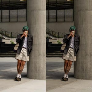 May include: A person wearing a black button-down shirt, beige shorts, and a green baseball cap. They are holding a white cup and standing next to a concrete pillar.