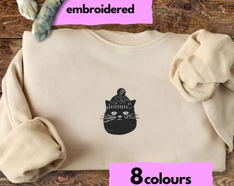 Op de afbeelding: Beige sweatshirt met een geborduurde zwarte kat die een hoed draagt. De tekst "embroidered" staat op een roze banner boven de sweatshirt. De tekst "8 colours" staat op een roze banner onder de sweatshirt. De tekst "Fur Family Studio Est 2024" staat op een witte cirkel met een zwarte omtrek.