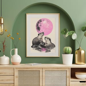 May include: A framed print of two capybaras looking at each other under a pink moon. The print is on a light beige background and is framed in a light wood frame. The print is hanging on a green wall above a wooden cabinet.