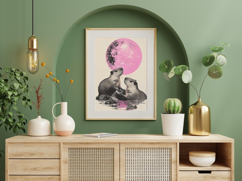 May include: A framed print of two capybaras looking at each other under a pink moon. The print is on a light beige background and is framed in a light wood frame. The print is hanging on a green wall above a wooden cabinet.