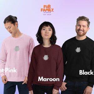 以下が含まれることがあります： 3色のスウェットシャツ。薄いピンク、マルーン、黒。各スウェットシャツには、小さなグレーの漫画の猫のグラフィックと、その上に「FUR FAMILY STUDIO EST 2024」というテキストが描かれています。