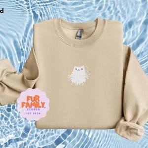 以下が含まれることがあります： 前面に白い刺繍の猫のデザインが施されたベージュのトレーナー。トレーナーはクルーネックで長袖です。画像の左上隅に「Sand」というテキストが表示されています。画像の左下隅のピンクの円の中に「Fur Family Studio Est 2024」というテキストが表示されています。