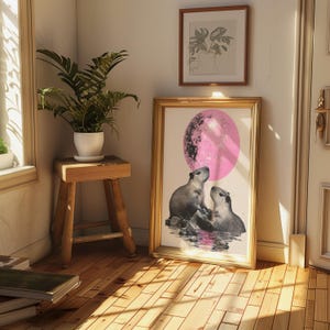 May include: A framed print of two capybaras looking up at a large pink moon. The capybaras are standing in a body of water with rocks and plants. The print is framed in gold.