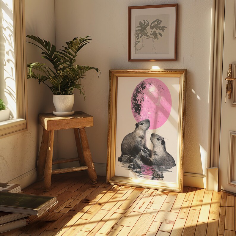 May include: A framed print of two capybaras looking up at a large pink moon. The capybaras are standing in a body of water with rocks and plants. The print is framed in gold.