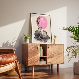 May include: A print of two capybaras sitting in a pool of water under a pink moon. The print is framed in a black frame and is hanging on a wall.
