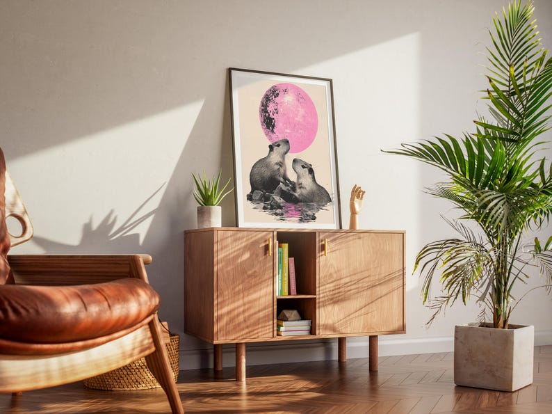May include: A print of two capybaras sitting in a pool of water under a pink moon. The print is framed in a black frame and is hanging on a wall.