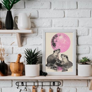 May include: A framed print of two capybaras looking up at a pink moon. The print is in a white frame and is hanging on a white brick wall.