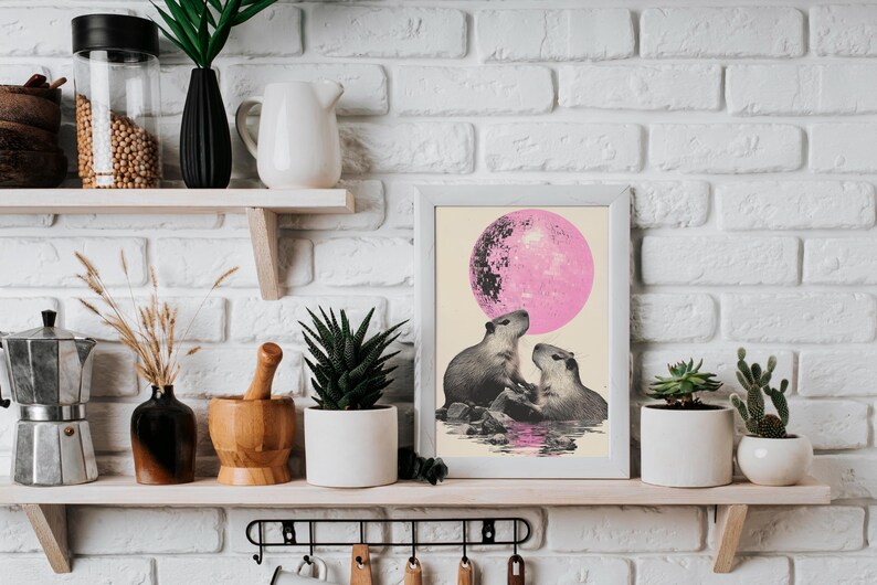 May include: A framed print of two capybaras looking up at a pink moon. The print is in a white frame and is hanging on a white brick wall.