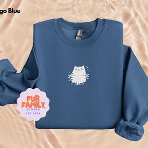 以下が含まれることがあります： インディゴブルーのトレーナーに、水たまりの中にいる猫の白い刺繍イラストが施されています。トレーナーには、ピンクの円の中に「Fur Family. Studio. Est 2024」という文字が書かれています。