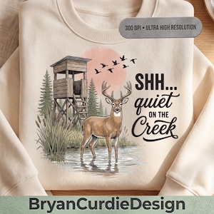 Pode incluir: Uma camisola de gola redonda cor de creme com uma impressão gráfica de um veado na água perto de um abrigo de caça de madeira, pinheiros e juncos. O texto diz SHH... quiet ON THE Creek, com um sol rosa e pássaros a voar acima.