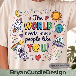 May include: A cream-colored t-shirt featuring a colorful graphic with the text The WORLD needs more people like YOU! surrounded by illustrations of a space shuttle, a smiling sun, a planet, an alien in a UFO, an astronaut, and stars.