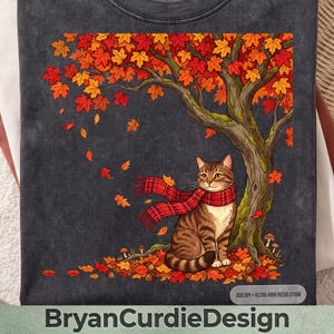 Puede incluir: Una camiseta gris carbón con un gráfico de un gato atigrado marrón con una bufanda de cuadros rojos, sentado bajo un gran árbol con hojas de otoño de color naranja y rojo. Hojas caídas y pequeñas setas cubren el suelo.