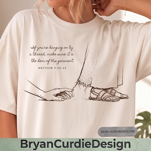 May include: A cream-colored t-shirt featuring a brown line-art illustration of a hand reaching for the hem of a garment. The text reads: If you're hanging on by a thread, make sure it's the hem of His garment. MATTHEW 9:20-22.