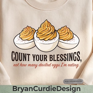 May include: A cream-colored crewneck sweatshirt featuring a graphic of three deviled eggs topped with paprika. Below the eggs, the text reads: COUNT YOUR BLESSINGS, not how many deviled eggs I'm eating. A small gray tag in the corner says 300 DPI ULTRA HIGH RESOLUTION.