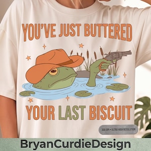 Peut inclure: Un t-shirt couleur crème avec un motif représentant une grenouille verte portant un chapeau de cowboy orange, émergeant de l'eau bleue, tenant un revolver, entourée de roseaux et d'étoiles. Le texte indique : YOU'VE JUST BUTTERED YOUR LAST BISCUIT.