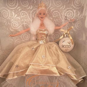 May include: A blonde doll wearing a gold gown with snowflake accents, a white fur stole, and a gold crown. The doll holds a Christmas ornament with the year 2000 on it. The doll is in a decorative box.