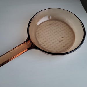 May include: Amber-colored glass frying pan with a long handle. The pan has a round cooking surface with a textured pattern. The rim and handle are a darker shade of amber.